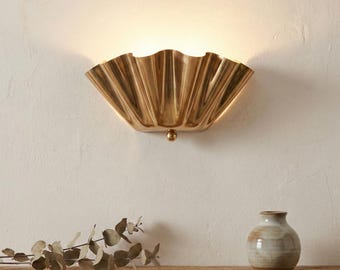 May include: A gold-toned metal wall sconce featuring a semi-circular, pleated fan design that radiates light upward. The fixture has a smooth, reflective surface and a small spherical accent at the bottom center, mounted against a plain wall.