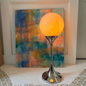 May include: A table lamp featuring a spherical, glowing orange glass shade supported by a slender, polished chrome metal stem and a wide, flared circular base. The lamp emits a warm light, standing upright on a flat surface.