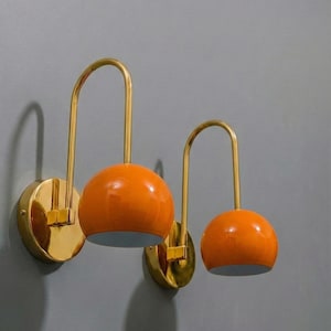 May include: Two wall sconces with a mid-century modern design. Each has a round, orange lampshade and a curved, gold-coloured arm. The sconces are mounted on a gold-coloured circular base.