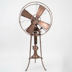 May include: A vintage copper-toned electric fan with four blades, a protective cage, and a decorative base. The fan stands on a tripod base and features an adjustable head. The overall design is reminiscent of early 20th-century industrial design.