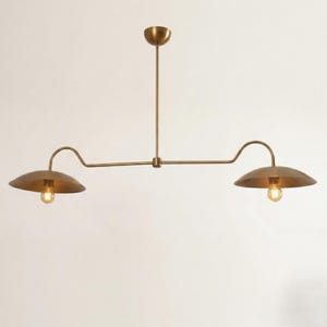 May include: A brass-toned metal chandelier featuring a central vertical rod connected to a horizontal bar with two curved arms. Each arm supports a wide, shallow, bowl-shaped shade with a single exposed light bulb pointing downward.