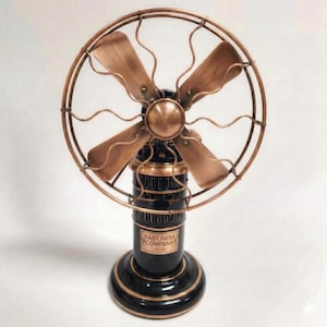 May include: A vintage-style electric fan with a black base and copper-coloured accents. The fan has four blades and a protective cage. The base features a plaque that reads "EAST INDIA COMPANY". A decorative piece for a vintage aesthetic.