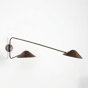 May include: A wall-mounted lamp featuring a dark bronze finish with two conical shades. The fixture has a long, slender, angular arm extending from a circular wall plate, supporting two downward-facing shades positioned at different heights.