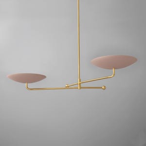 Puede incluir: Un candelabro moderno y minimalista con acabado de latón. Dos pantallas en forma de platillo, de color rosa rubor, están suspendidas de brazos curvos. La lámpara cuelga de una varilla central, creando un diseño equilibrado y elegante.