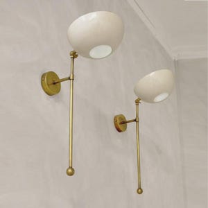 Puede incluir: Un par de lámparas de pared con pantallas en forma de cuenco de color blanco roto unidas a brazos largos y delgados de latón. Cada lámpara tiene una placa de pared circular de latón y una pequeña esfera de latón en la base de la varilla.