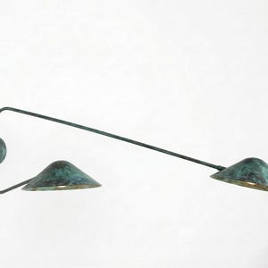 May include: A wall-mounted, two-light fixture with a weathered, verdigris finish. The fixture has a circular base, a curved arm, and two conical shades. The shades are angled downward, and the fixture is mounted on a white wall.