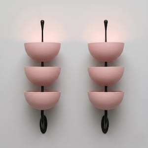 Puede incluir: Un par de apliques de pared, cada uno con tres cuencos semiesféricos de color rosa mate apilados sobre una varilla vertical negra central. El cuenco superior de cada lámpara emite un brillo cálido y suave, mientras que la varilla negra se extiende por encima y por debajo.