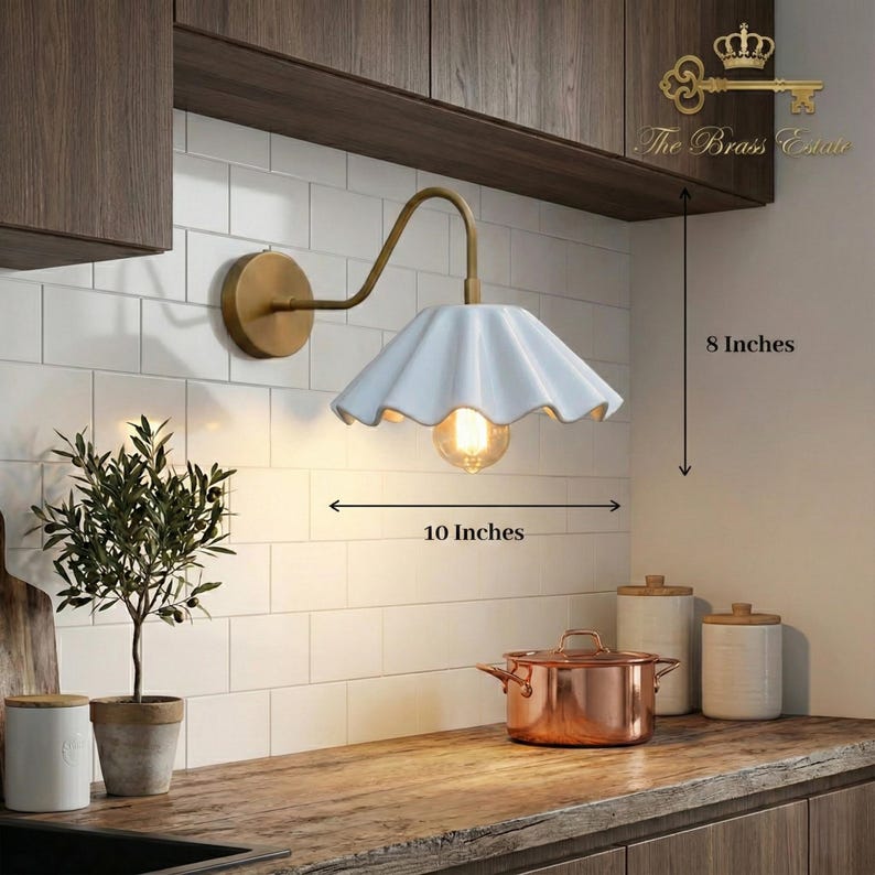 May include: A wall-mounted lamp with a brass-colored arm and a white, fluted, scalloped ceramic shade. The lamp features a visible light bulb and includes text indicating dimensions of 8 inches in height and 10 inches in width.