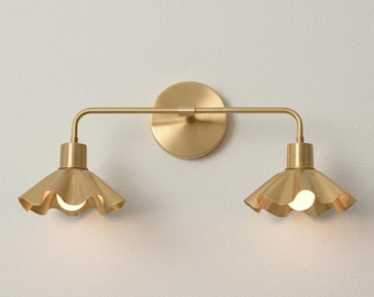May include: A two-light wall sconce featuring a brushed brass finish. The fixture has a circular wall plate, a horizontal bar, and two downward-facing shades with a scalloped, ruffled edge design. Each shade contains a single light bulb.
