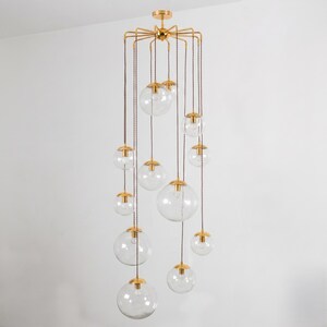 May include: A multi-light chandelier featuring a gold-toned metal frame with twelve arms extending from a central hub. Each arm supports a clear glass globe pendant suspended by a brown twisted fabric cord at varying lengths from the ceiling.