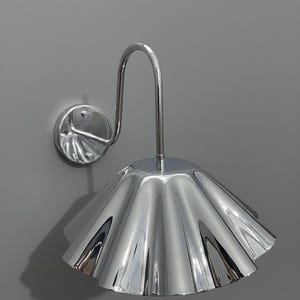 May include: A wall-mounted lamp featuring a polished chrome finish with a reflective, fluted, bell-shaped shade. The fixture includes a curved metal arm extending from a circular base plate, creating a sleek, metallic appearance with visible light reflections.
