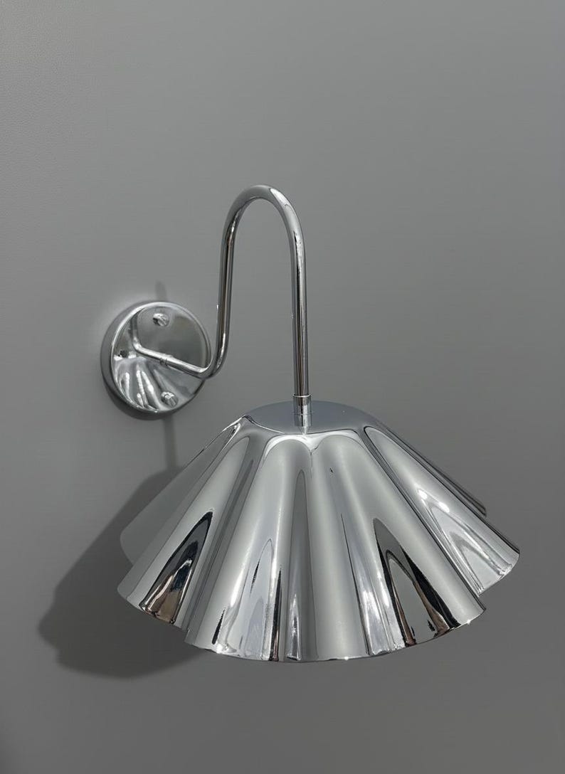 May include: A wall-mounted lamp featuring a polished chrome finish with a reflective, fluted, bell-shaped shade. The fixture includes a curved metal arm extending from a circular base plate, creating a sleek, metallic appearance with visible light reflections.