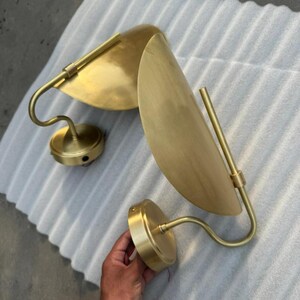 May include: Two wall sconces featuring a brushed brass finish. Each fixture consists of a circular wall-mounting plate, a curved arm, and a semi-circular shade that directs light. The metal surfaces have a smooth, matte, golden-toned appearance.