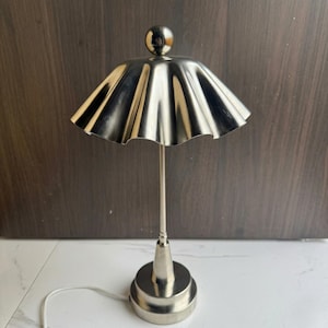 May include: A silver-toned table lamp with a ruffled shade and a spherical finial. The lamp has a slender stem and a circular base. The cord is white. The lamp is approximately 12 inches tall.