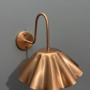May include: A wall-mounted lamp featuring a metallic copper finish. The fixture consists of a circular wall plate, a curved arm, and a wide, fluted, bell-shaped shade with a scalloped edge that directs light downward from the central stem.