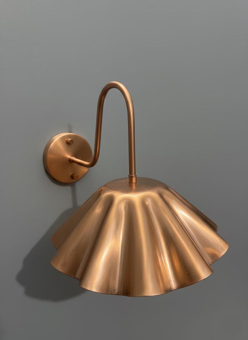 May include: A wall-mounted lamp featuring a metallic copper finish. The fixture consists of a circular wall plate, a curved arm, and a wide, fluted, bell-shaped shade with a scalloped edge that directs light downward from the central stem.
