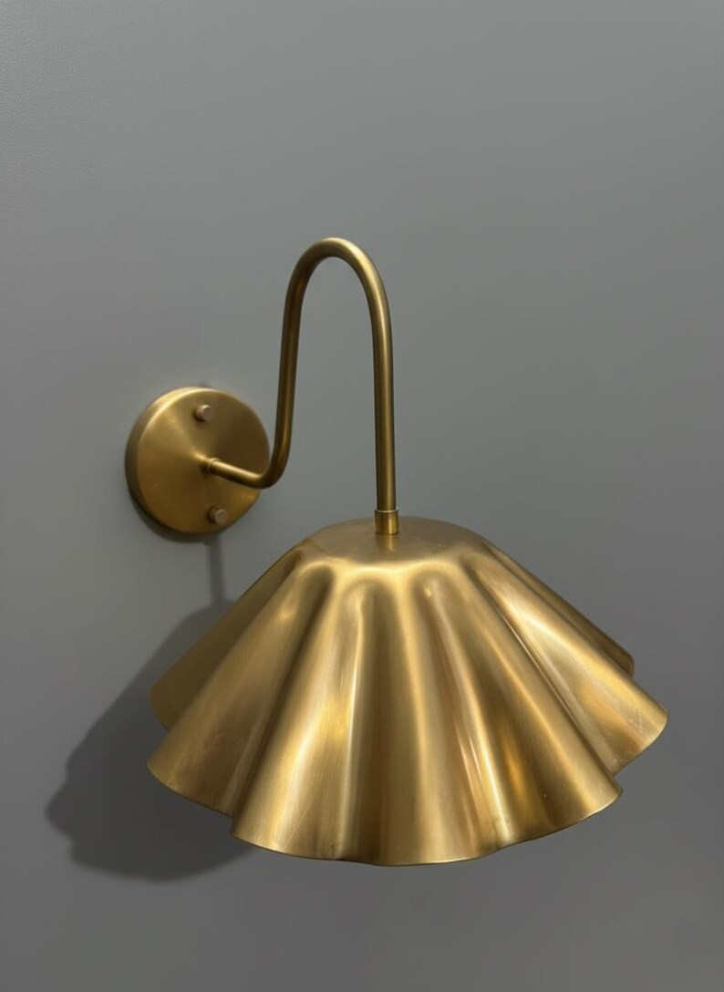 May include: A wall-mounted lamp featuring a brushed brass finish. The fixture includes a circular mounting plate, a curved arm extending outward, and a wide, fluted, scalloped-edge shade that directs light downward in a bell-like shape.
