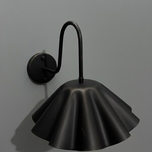May include: A matte black wall sconce featuring a curved metal arm attached to a circular wall plate. The light fixture includes a wide, downward-facing shade with a distinctively ruffled, pleated edge, creating a scalloped silhouette.