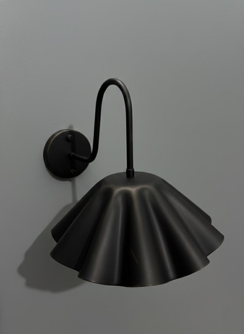 May include: A matte black wall sconce featuring a curved metal arm attached to a circular wall plate. The light fixture includes a wide, downward-facing shade with a distinctively ruffled, pleated edge, creating a scalloped silhouette.