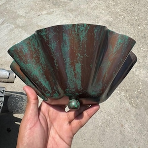 May include: A semi-circular, fan-shaped metal bowl featuring a dark brown base color with a distressed, weathered teal green patina finish. The bowl has a pleated, fluted design and a small, spherical knob at the bottom center point.