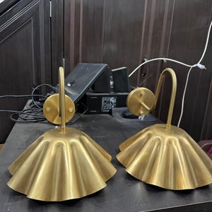 May include: Two matching wall sconces featuring a brushed gold finish. Each lamp has a circular wall-mounting plate, a curved arm, and a wide, pleated, bell-shaped shade that flares outward at the bottom edge, creating a distinct rippled texture.