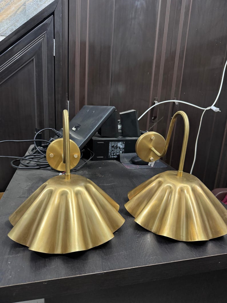 May include: Two matching wall sconces featuring a brushed gold finish. Each lamp has a circular wall-mounting plate, a curved arm, and a wide, pleated, bell-shaped shade that flares outward at the bottom edge, creating a distinct rippled texture.