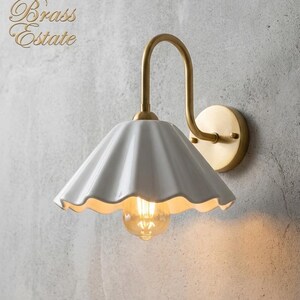 wall light with a white, scalloped lampshade and a brass-coloured arm and base. The light bulb is visible, emitting a warm glow. The fixture is mounted on a textured, grey wall. The Brass Estate is written in