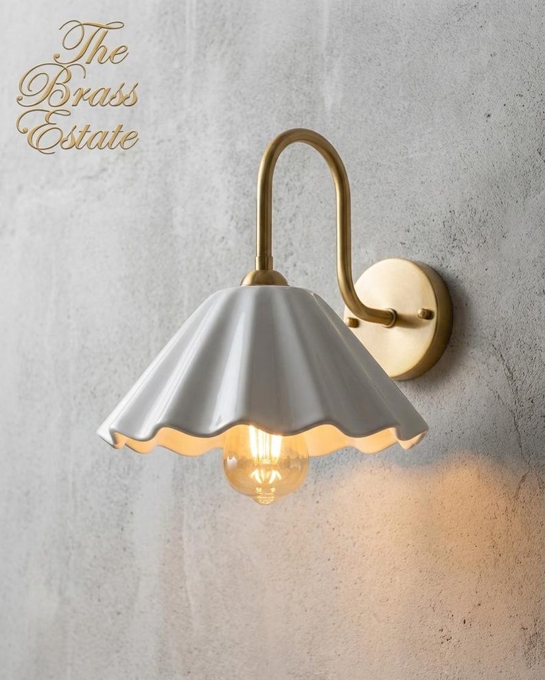 wall light with a white, scalloped lampshade and a brass-coloured arm and base. The light bulb is visible, emitting a warm glow. The fixture is mounted on a textured, grey wall. The Brass Estate is written in