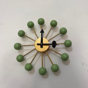 May include: A mid-century modern wall clock with a gold-colored face and hands. The clock features gold-colored spokes with green spherical accents. The clock's hands are black.