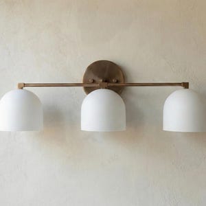 May include: A three-light wall fixture featuring a horizontal brass-toned metal bar and a circular mounting plate. Three matte white dome-shaped glass shades are evenly spaced along the bar, providing a clean and symmetrical lighting design.