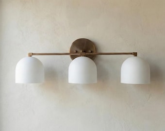 May include: A three-light wall fixture featuring a horizontal brass-toned metal bar and a circular mounting plate. Three matte white dome-shaped glass shades are evenly spaced along the bar, providing a clean and symmetrical lighting design.