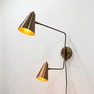 May include: A dual-arm wall lamp featuring a metallic brass finish with two cone-shaped shades. The fixture is mounted to a circular wall plate and includes two adjustable arms, each ending in a conical light head emitting a warm glow.