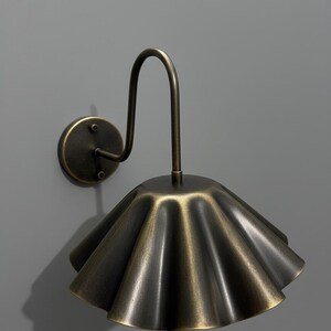 May include: A wall-mounted lamp featuring a dark brass finish with a curved arm and a wide, pleated, fan-shaped shade. The metal surface has a brushed, aged texture, and the fixture is attached to a circular base plate with two visible screws.