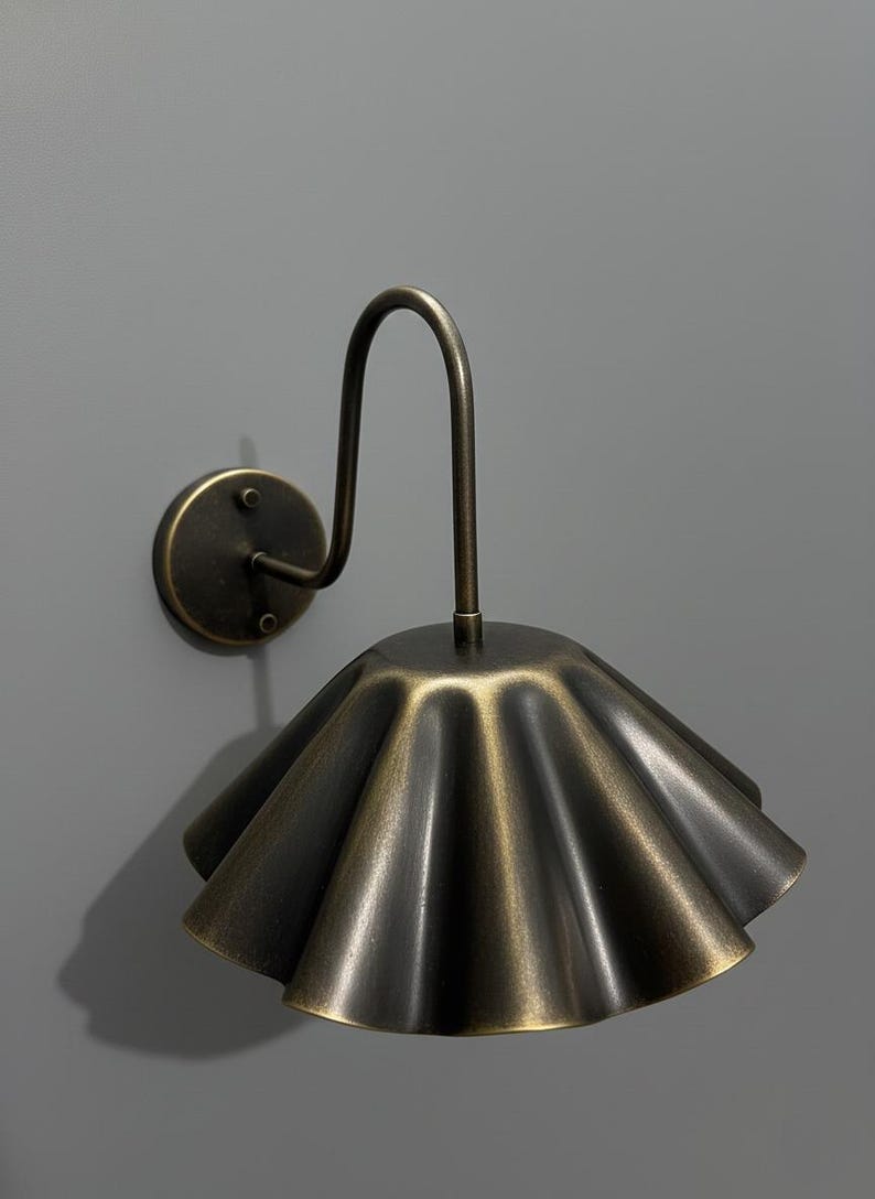 May include: A wall-mounted lamp featuring a dark brass finish with a curved arm and a wide, pleated, fan-shaped shade. The metal surface has a brushed, aged texture, and the fixture is attached to a circular base plate with two visible screws.