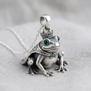 May include: A silver frog pendant with a crown and green gemstone eyes. The frog is wearing a cape and a small green bow tie. The pendant hangs from a delicate silver chain.