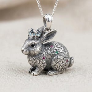 Könnte beinhalten: Ein Kaninchen-Anhänger aus Sterlingsilber mit einer Krone aus hellblauem Emaille und kleinen klaren Steinen. Der Körper des Kaninchens ist mit aufwendigen Gravuren im Rankenmuster verziert und mit kleinen roten und grünen Edelsteinen besetzt.