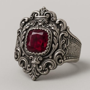 Könnte beinhalten: Ein silberfarbener Ring mit einem kunstvollen, verschnörkelten Design. Er hat einen großen, quadratisch geschliffenen, tiefroten Edelstein. Das Band ist gemustert, was den Vintage-Stil des Rings unterstreicht.