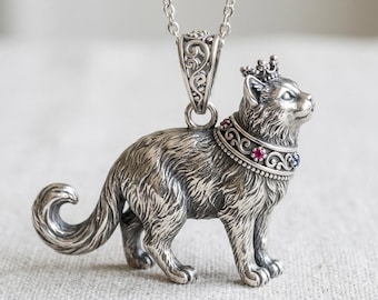 May include: A silver cat pendant with a crown and a decorative collar featuring red and blue gemstones. The cat is standing and has a curled tail. The pendant hangs from a silver chain.