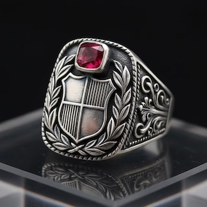 May include: A silver ring with a shield design and a red gemstone. The ring features a detailed crest with a laurel wreath and ornate scrollwork on the sides. The gemstone is square-cut.