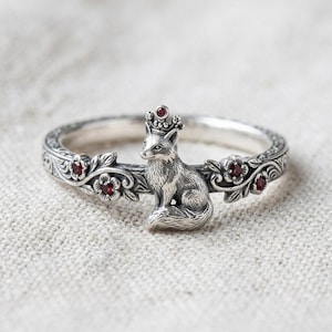 May include: A silver ring featuring a sculpted fox wearing a small crown. The band is decorated with embossed floral vines and set with four small, round, deep red gemstones. The metal has an oxidised finish to highlight the intricate details.