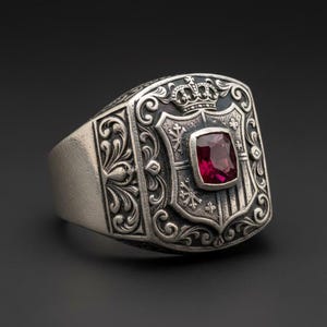 May include: Silver ring with a shield design, a crown, and a rectangular red gemstone. The ring has intricate scrollwork and a vintage aesthetic. The gemstone is approximately 0.25 inches in size.