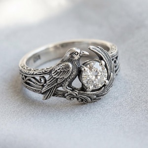 May include: A silver ring featuring a detailed raven perched on a branch, holding a large, clear gemstone. The ring has an ornate, engraved band with a nature-inspired design. The gemstone is round and faceted, catching the light.