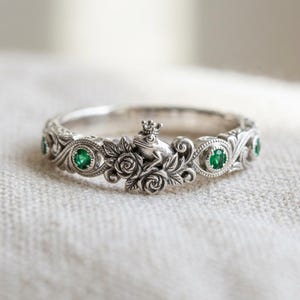 May include: A silver ring featuring a central frog wearing a crown, surrounded by sculpted roses and leaves. The band is decorated with intricate scrollwork and set with small, round, vibrant green gemstones on either side of the central motif.