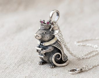 May include: A silver pendant necklace featuring a detailed mouse wearing a crown with red gemstones. The mouse is also wearing a cape and holding a scroll. The pendant hangs from a delicate silver chain.