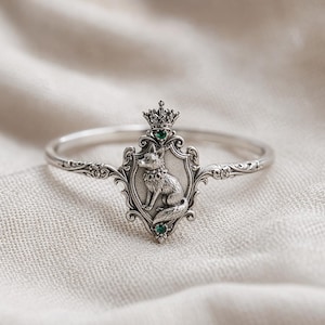 May include: Silver-toned bracelet featuring a detailed fox emblem with a crown and green gemstones. The fox is centered on a decorative shield, with ornate scrollwork on the bracelet's band. The bracelet is set against a neutral fabric backdrop.