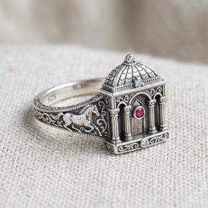Op de afbeelding: Een ring van sterling zilver met een gedetailleerde architecturale koepelstructuur met zuilen, een kleine ronde rode edelsteen en kleine heldere steentjes. De ringband toont een reliëfmotief van een rennend paard met gedetailleerd krulwerk.