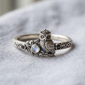 Könnte beinhalten: Ein silberner Ring mit einer detaillierten Eule, die eine kleine Krone trägt und auf einem Ast sitzt. Der Ring ist mit kunstvollen Schnörkeln, einem runden, schillernden Mondstein und zwei kleinen schwarzen, runden Edelsteinen verziert.