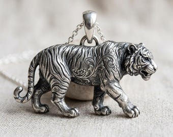 May include: A silver tiger pendant with intricate floral and stripe detailing. The pendant is attached to a delicate chain. The tiger is depicted in a walking pose, showcasing its detailed features. The pendant is likely made of sterling silver.