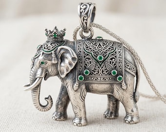 May include: A silver elephant pendant with intricate detailing. The elephant wears a crown and is adorned with green gemstones. The pendant hangs from a silver chain, showcasing a detailed design.