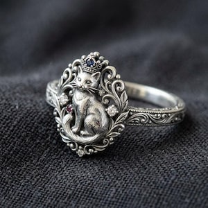 May include: A silver ring featuring a detailed cat design wearing a crown, set within an ornate oval frame. The ring is embellished with small gemstones and intricate scrollwork. The ring is on a dark gray surface.